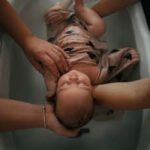 4 mains tenant un bébé enveloppé dans un lange dans une baignoire d'eau pour un rituel du bain sensoriel