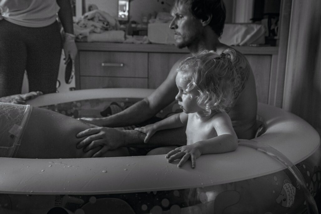 enfant et son père dans une piscine d'accouchement. les mains sur le bas du dos de la future maman. Une personne à l'arrière plan est présente à l'accouchement